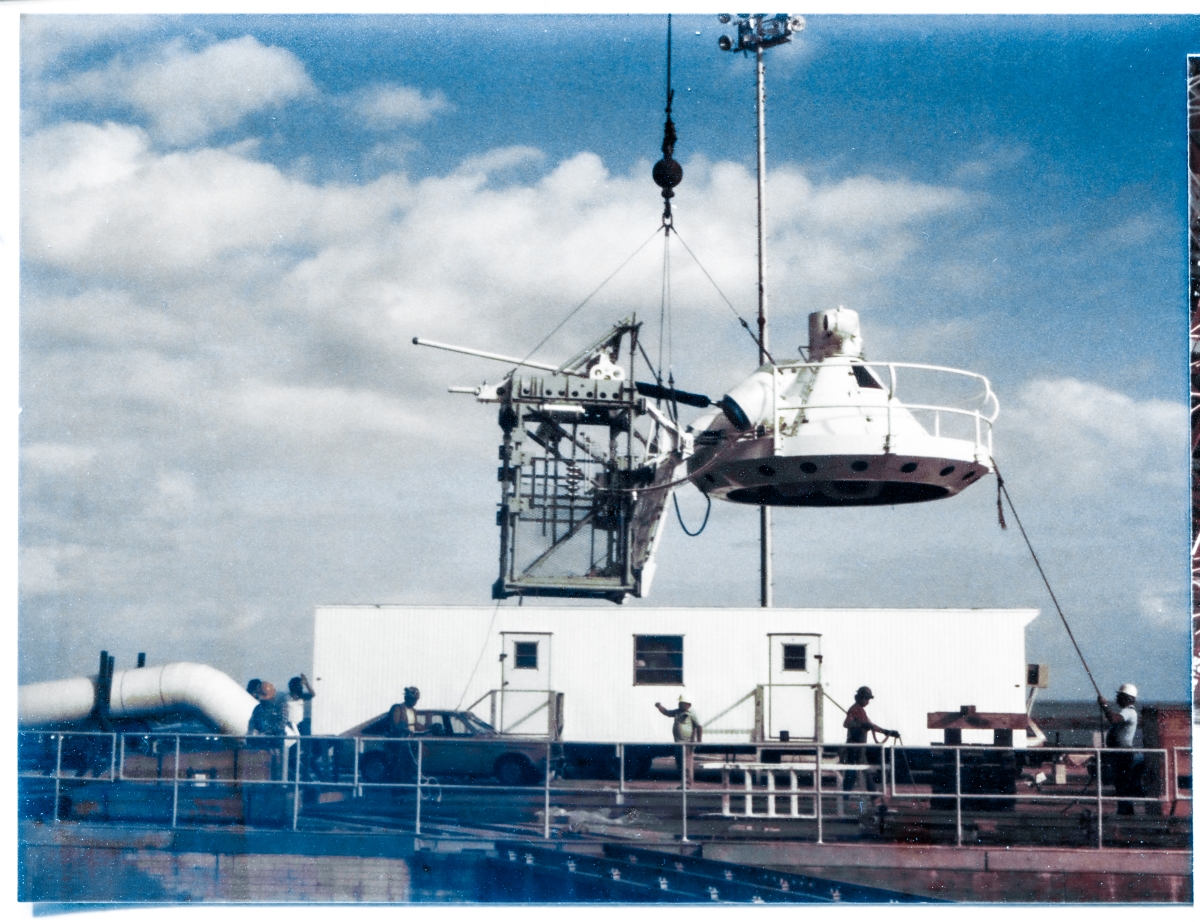 Image 117. At Space Shuttle Launch Complex 39-B, Kennedy Space Center, Florida, the External Tank Gaseous Oxygen Vent System Tip Assembly, which included the gray steel framework that held all of the mechanical components for lifting, lowering, and orienting the white flying-saucer-shaped Gaseous Oxygen Vent Hood which you see attached to the right side of that steel framework, more commonly known as the ”Beanie Cap”, rises from the Pad Deck where it was originally delivered to and stored, prior to being lifted and bolted into place on the end of the GOX Arm near the top of the Fixed Service Structure, by Union Ironworkers working for Ivey Steel. Photo by James MacLaren.